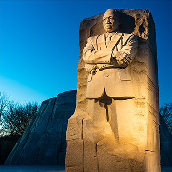 Statue of Martin Luther King Jr., carved roughly out of stone. His gaze stern and intent, arms crossed across his chest.